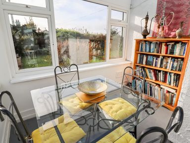 A dining area with a glass table and yellow cushioned chairs next to a bookshelf filled with books at The Flat At Brwyn in Brynford