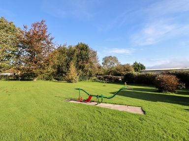 A grassy outdoor area with a green and red farming plow on a paved section and trees in the background at The Flat At Brwyn in Brynford
