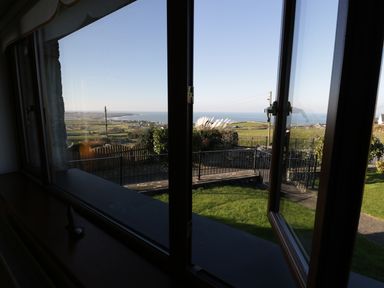 A view through an open window showing a garden with grass and a fence with the sea and coastline in the distance at Awel Y Llan in Llanfair