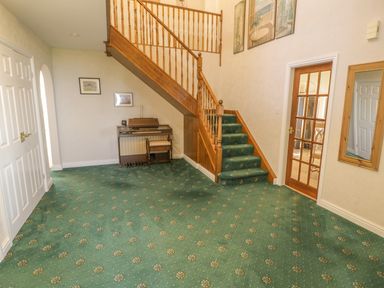 A hallway with a green carpeted staircase wooden banister an electronic organ two framed pictures and a glass panel door at Awel Y Llan in Llanfair