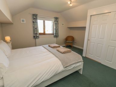 A bedroom with a bed with white sheets and towels on it a wicker chair a window with curtains and a white closet at Awel Y Llan in Llanfair
