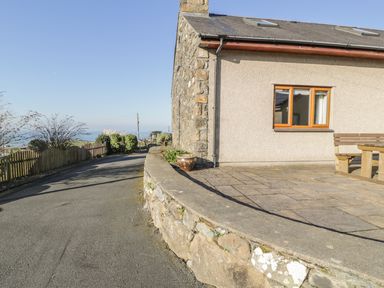 An exterior view of a stone and stucco house with a paved patio and wooden bench beside a curved road at Awel Y Llan in Llanfair