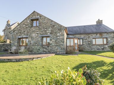 A stone house with wooden framed windows and a stone patio with chairs surrounded by grass at Awel Y Llan in Llanfair