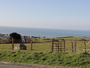 A grassy field with a wire fence and gate overlooking houses and the ocean at Awel Y Llan in Llanfair