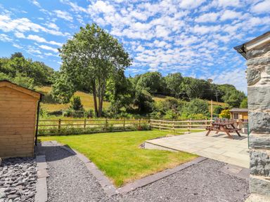 A backyard with a wooden shed a tree picnic table and a stone patio at Ffrwdd Galed Uchaf in Tregarth