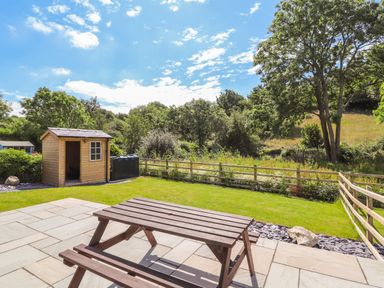 A wooden picnic table on a stone patio overlooking a fenced grassy backyard with a wooden shed and trees at Ffrwdd Galed Uchaf in Tregarth