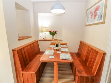 A dining nook with wooden benches and table set with placemats and glasses a bottle and a lamp with flowers on a ledge and a framed picture of chickens on the wall at Ffrwdd Galed Uchaf in Tregarth