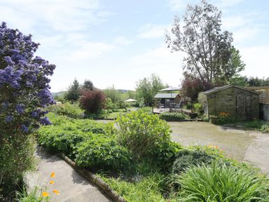 A garden with a stone path shrubs trees a wooden shed and patio furniture with umbrellas at Rhydygaled in Llanrhaeadr-Ym-Mochnant
