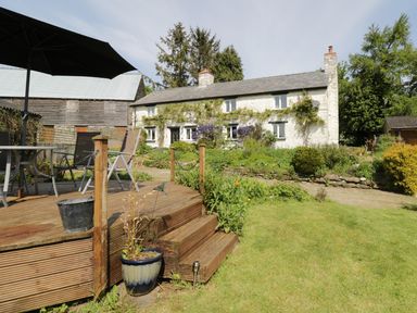 A garden with a wooden deck patio and a stone house with plants and trees at Rhydygaled in Llanrhaeadr-Ym-Mochnant