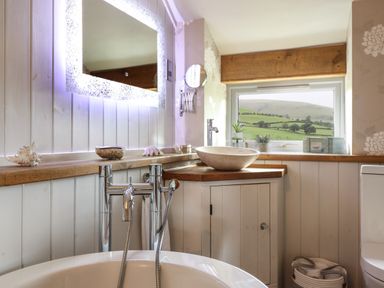 A bathroom with a freestanding bathtub a lit mirror a countertop basin and a window showing green hills at Rhydygaled in Llanrhaeadr-Ym-Mochnant