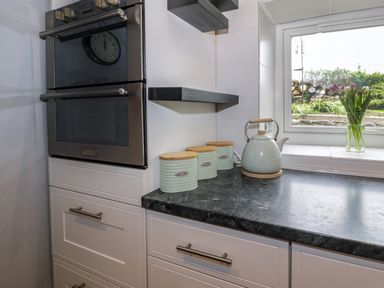 A kitchen corner with white cabinets black countertop an oven three storage canisters a kettle and a window with flowers on the sill at Rhydygaled in Llanrhaeadr-Ym-Mochnant