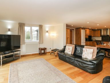A living room with a black leather sofa a large TV on a stand a rug wooden floor and a view of a kitchen with wooden cabinets at The Annex in Royal Wootton Bassett