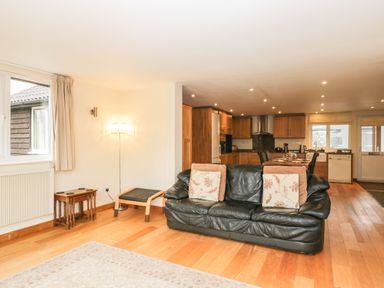 A living room with a black leather sofa and wooden tables next to a kitchen area at The Annex in Royal Wootton Bassett