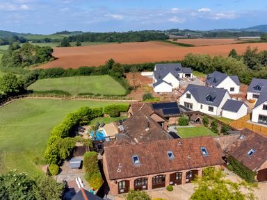 Aerial view of houses with brown and gray roofs surrounding a swimming pool and green fields at The Old Byres in Washford near Watchet