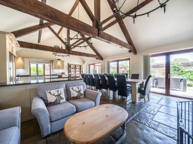 An open plan room with wooden beams a sofa with cushions a wooden coffee table and a large dining table with chairs at The Old Byres in Washford near Watchet