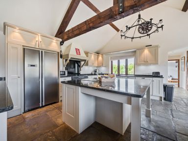 A kitchen with an island, stainless steel refrigerator, stove, cabinets, stone floor, and exposed wooden beams at The Old Byres in Washford near Watchet