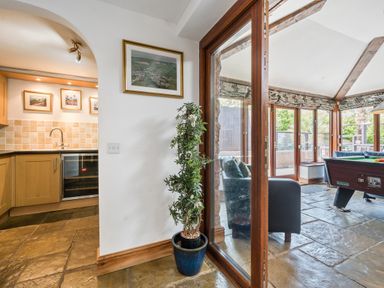 A kitchen with wooden cabinets and a sink next to a potted plant near glass doors leading to a room with a pool table and armchairs at The Old Byres in Washford near Watchet