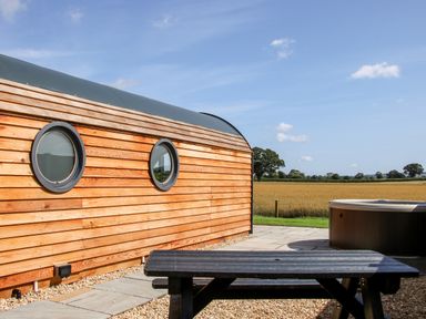 A wooden cabin with round windows beside a patio with a bench and a hot tub overlooking a field at Withy Willows in Diddlebury near Ludlow