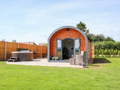 A wooden pod with open doors on a patio with chairs and a hot tub in a fenced garden at Withy Willows in Diddlebury near Ludlow
