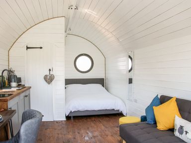 A small room with a bed a sofa with colorful cushions a round window a wooden floor and a kitchenette at Withy Willows in Diddlebury near Ludlow