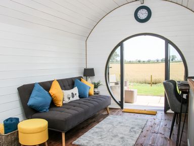 A room with a dark gray sofa with blue and yellow cushions a yellow stool a round glass door overlooking a field and a clock on the wall at Withy Willows in Diddlebury near Ludlow