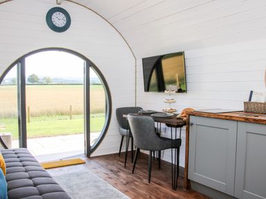 A room with a round door opening to a field a wall clock a wall-mounted television a table with chairs and plates and a kitchen cabinet at Withy Willows in Diddlebury near Ludlow