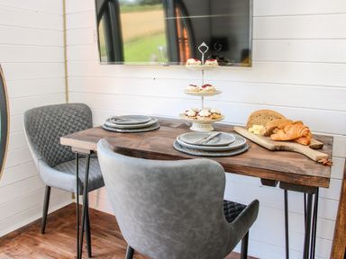 A dining table set with plates cups and cutlery with pastries and bread on a wooden board in a small room with grey chairs and a wall mounted tv at Withy Willows in Diddlebury near Ludlow