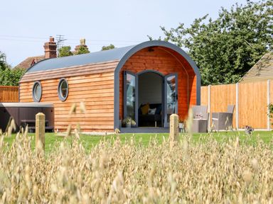 A wooden pod with a curved roof and glass doors opening onto a lawn with outdoor chairs and a hot tub at Withy Willows in Diddlebury near Ludlow