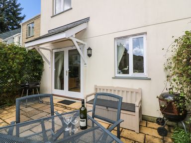 A patio with a metal table and chairs a storage bench barbecue grill and a glass door and window at Oyster Cottage in Falmouth