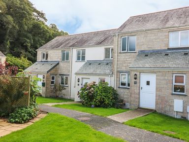 A row of stone and brick terraced houses with white doors and windows along a paved path with green lawns and shrubs Oyster Cottage in Falmouth
