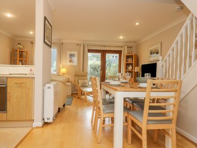 A dining area with a wooden table and chairs set with plates and glasses next to a living room with armchairs and a bookshelf at Oyster Cottage in Falmouth