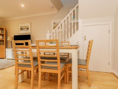 A dining area with a wooden table and six chairs near a white staircase and a bookcase in a living room at Oyster Cottage in Falmouth