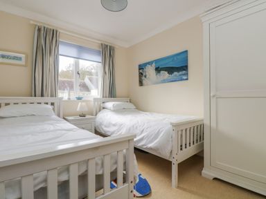 A bedroom with two single white beds a white nightstand with a lamp a window with curtains a white wardrobe and a seascape picture on the wall at Oyster Cottage in Falmouth