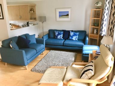 A living room with blue sofas a cream armchair a wooden shelving unit and a kitchen pass-through at Oyster Cottage in Falmouth