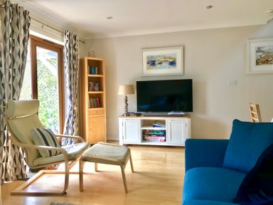 A living room with a blue sofa a beige armchair and ottoman a wooden bookshelf and a TV on a white stand at Oyster Cottage in Falmouth