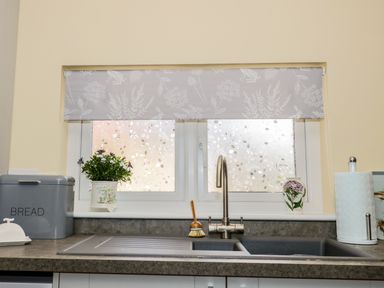 A kitchen sink with a faucet and brush with plants on the windowsill and a bread box and paper towel holder at Woodpecker Lodge in Henley near Langport