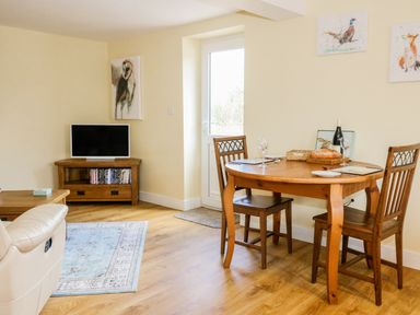 A room with a wooden dining table, two chairs, a small TV on a wooden stand, and animal paintings on the wall at Woodpecker Lodge Henley near Langport