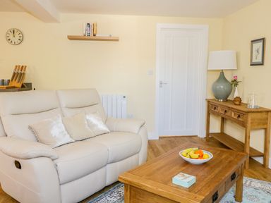 A living room with a cream sofa and cushions a wooden coffee table with fruit a console table with a lamp and vase and a wall shelf with books at Woodpecker Lodge in Henley near Langport