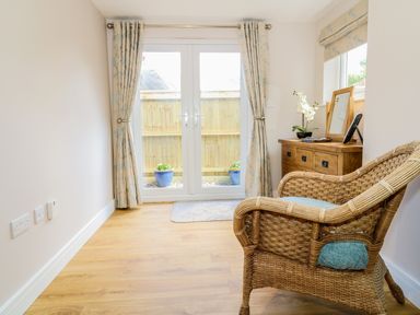 A room with a wicker chair wooden dresser with a mirror and double glass doors with curtains at Woodpecker Lodge in Henley near Langport
