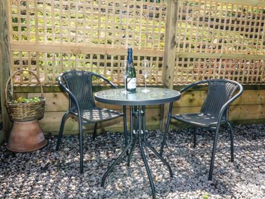 A small round glass table with two chairs on gravel and a bottle with two glasses on the table at Woodpecker Lodge in Henley near Langport