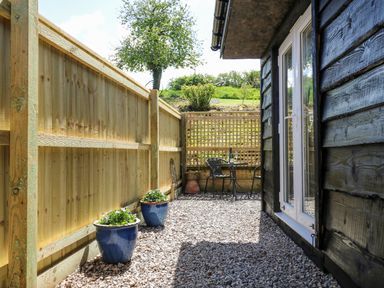 A narrow outdoor patio area with gravel ground two blue plant pots wooden fence and small table with two chairs at Woodpecker Lodge in Henley near Langport