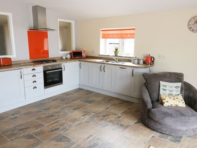 A kitchen with white cabinets and orange appliances next to a grey armchair with cushions at Knott Lodge in Sedbergh
