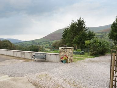 An outdoor gravel area with a bench a stone pillar with flower pots trees and hills in the background at Knott Lodge in Sedbergh