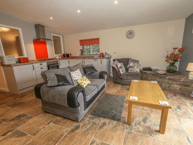 A living room with a gray sofa and armchair a wooden coffee table and kitchen area with white cabinets at Knott Lodge in Sedbergh