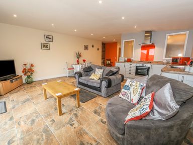 An open living room and kitchen with sofas, a wooden coffee table, a dining table with chairs, and kitchen appliances at Knott Lodge in Sedbergh