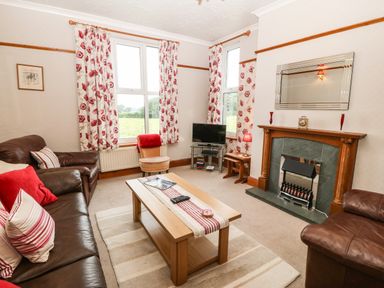 A living room with two windows floral curtains a glass TV stand a wooden coffee table and a fireplace at Dinas Farmhouse Annex in Caernarfon