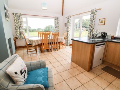 A kitchen dining area with wooden table and chairs next to large windows and a kitchen counter with appliances at Dinas Farmhouse Annex in Caernarfon