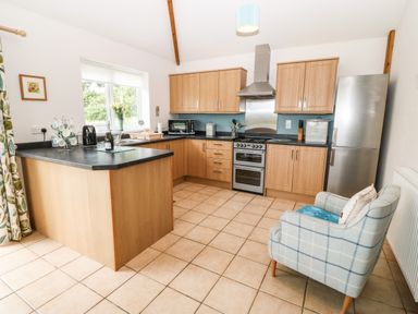 A kitchen with wooden cabinets a stove refrigerator a countertop with a kettle and cups and a checkered armchair near a radiator at Dinas Farmhouse Annex in Caernarfon
