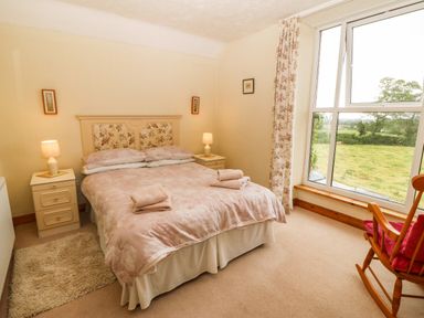 A bedroom with a double bed two bedside tables with lamps a rocking chair and a large window showing a garden at Dinas Farmhouse Annex in Caernarfon