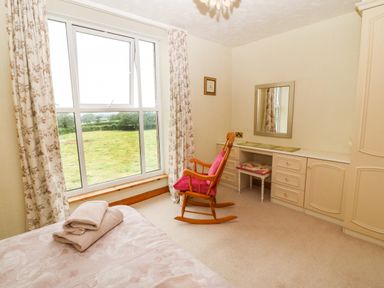 A bedroom with a bed, folded towels, a window with floral curtains, a rock chair with pink cushions, a vanity desk with mirror and a cushioned stool at Dinas Farmhouse Annex Caernarfon